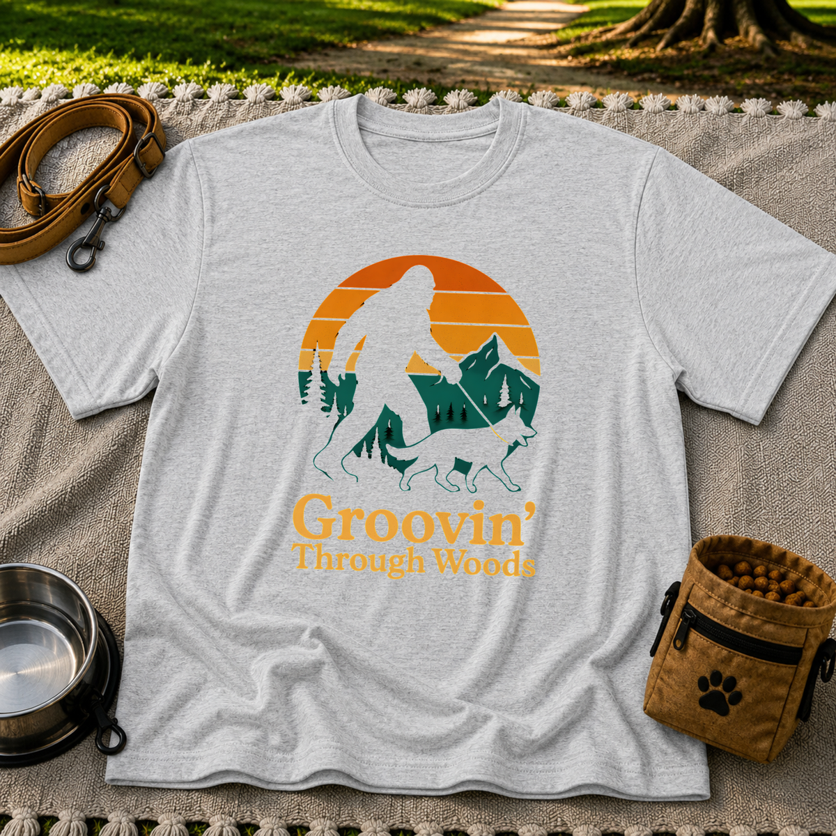 Sport Grey T-shirt mockup featuring Bigfoot and a dog walking through pine trees beneath an orange sunset.