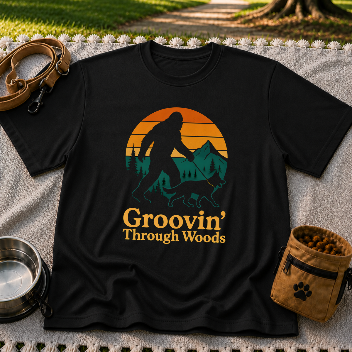 Black T-shirt mockup featuring Bigfoot and a dog walking through pine trees beneath an orange sunset.