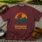 Maroon T-shirt mockup featuring Bigfoot and a dog walking through pine trees beneath an orange sunset.