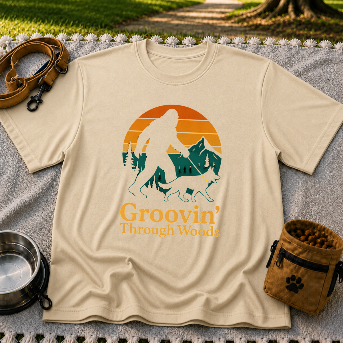 Sand T-shirt mockup featuring Bigfoot and a dog walking through pine trees beneath an orange sunset.