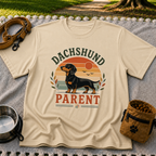 Sand T-shirt mockup featuring dachshund portrait in a striped sunset badge with Dachshund Parent lettering.
