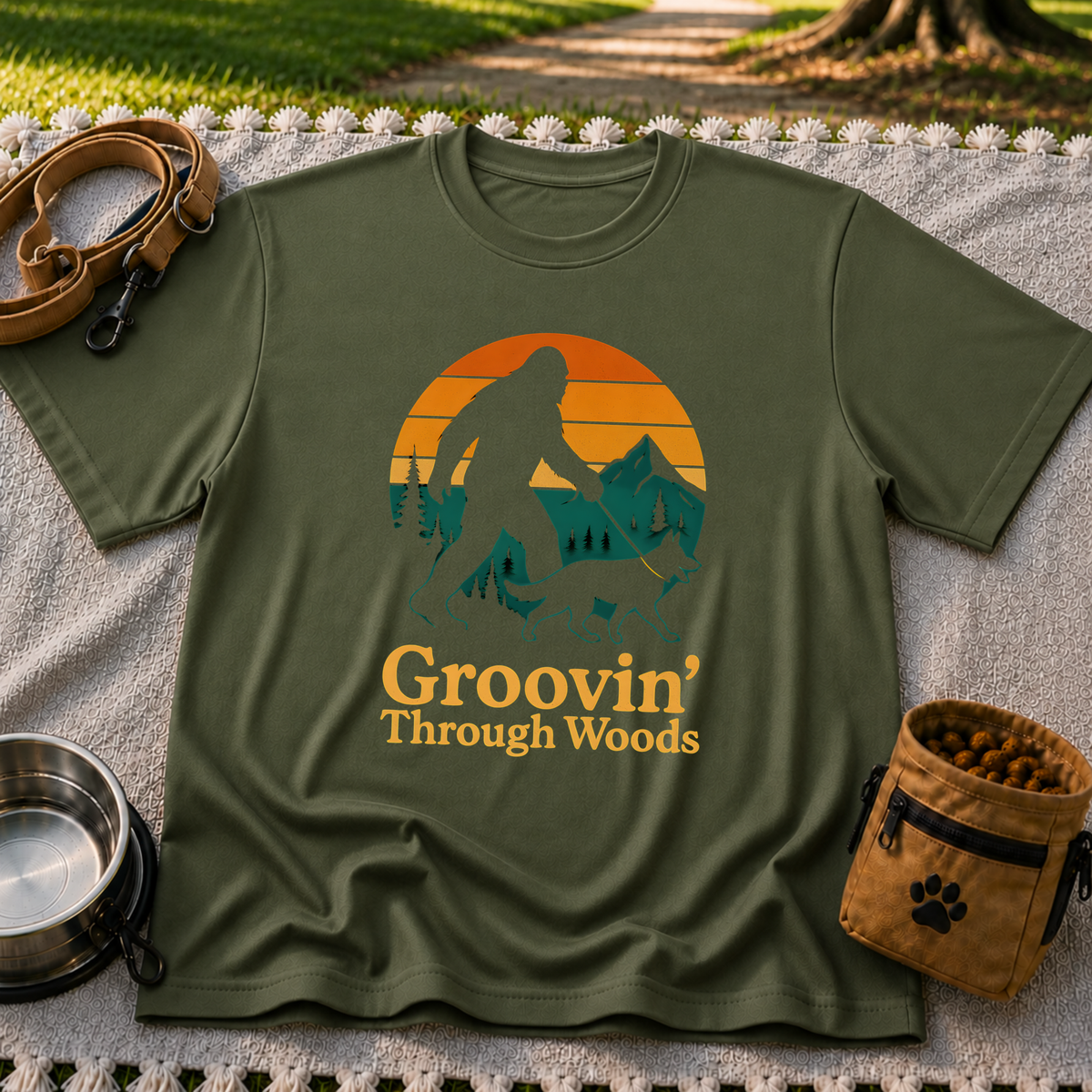 Military Green T-shirt mockup featuring Bigfoot and a dog walking through pine trees beneath an orange sunset.
